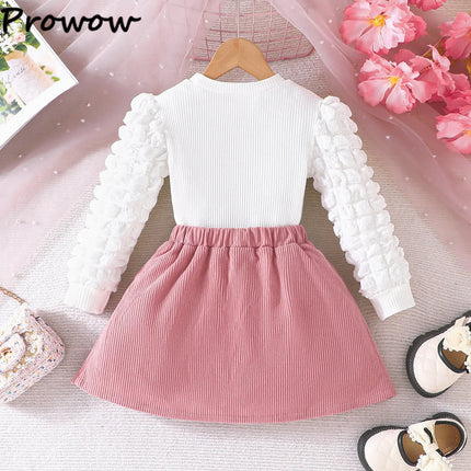 White long-sleeve top with ruffled sleeves and a pink skirt on a hanger against a decorative background.

